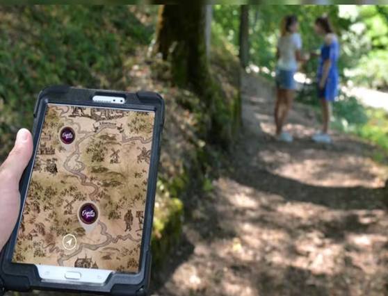 Tablette tenue en main affichant une carte au trésor interactive, avec deux personnes sur un sentier en forêt en arrière-plan