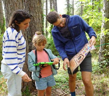 Une mère, une enfant et un père en forêt examinant une tablette et une planche d'énigmes en bois