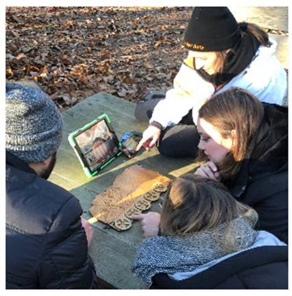 Une famille en cercle autour d une tablette interactive et d une planche d énigmes en bois sur une table en forêt